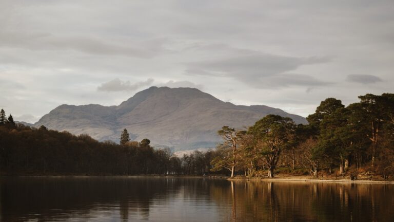 A lake, trees and mountains under a cloudy Scottish sky.
