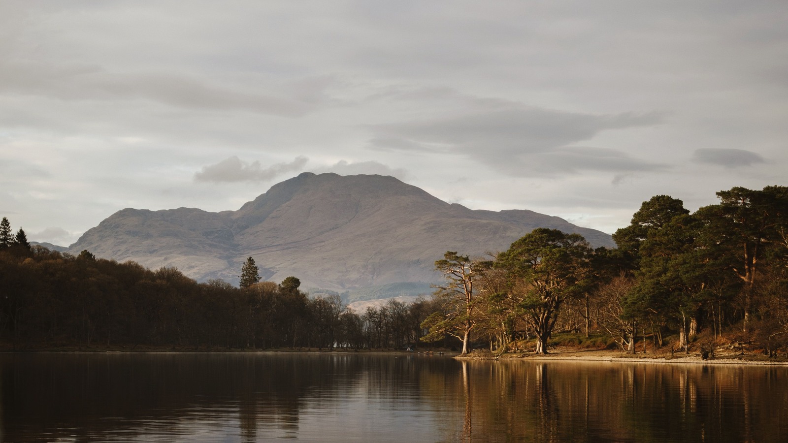 A lake, trees and mountains under a cloudy Scottish sky.