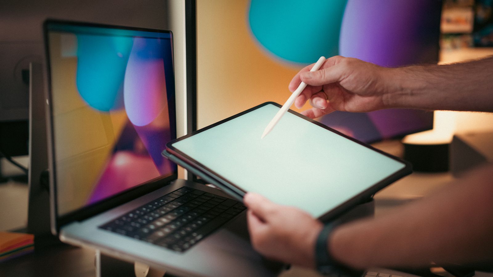 Hands holding a tablet and stylus beside a laptop and monitor.