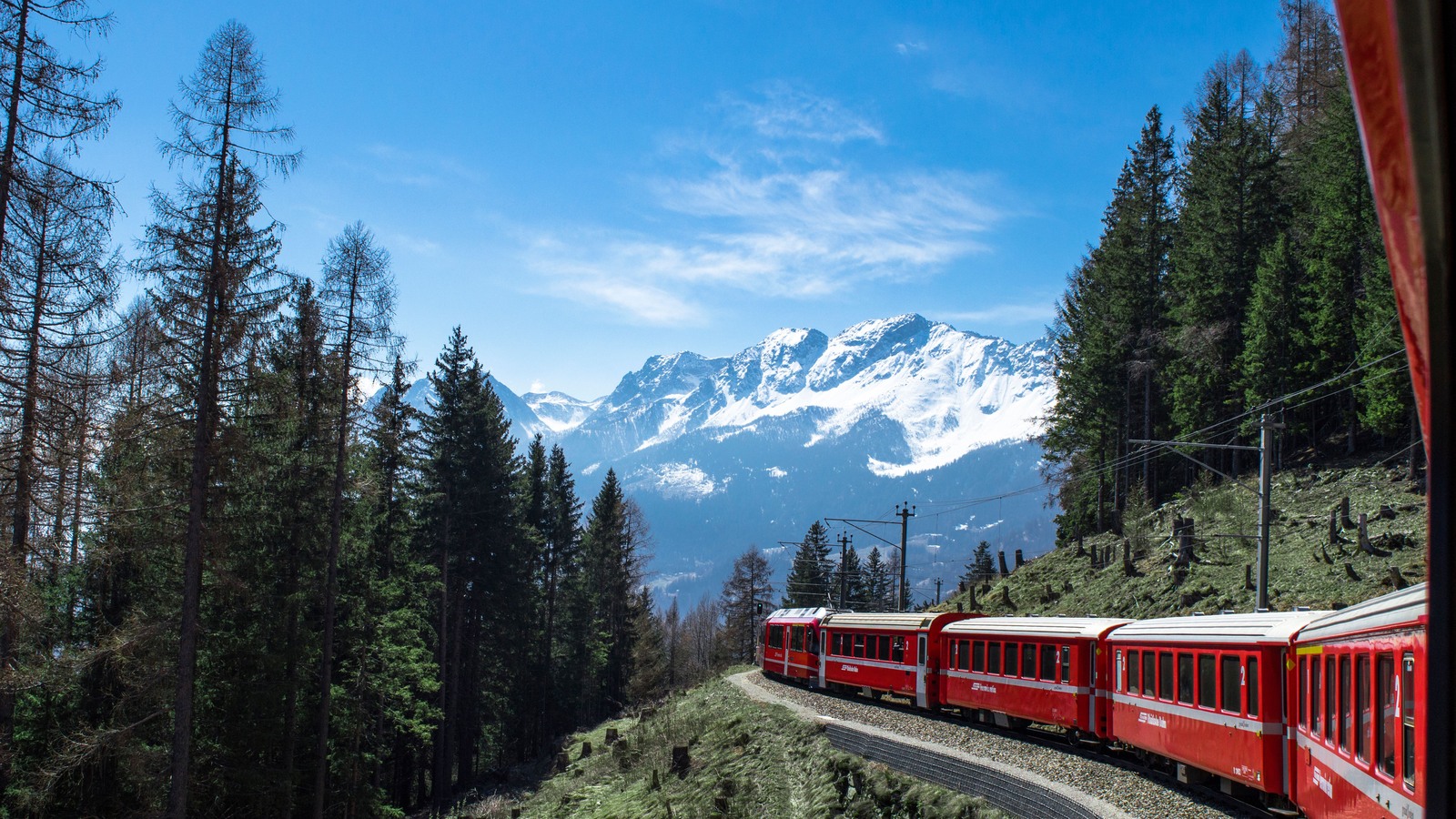 A red train curves through forest beneath snowy Swiss mountains.